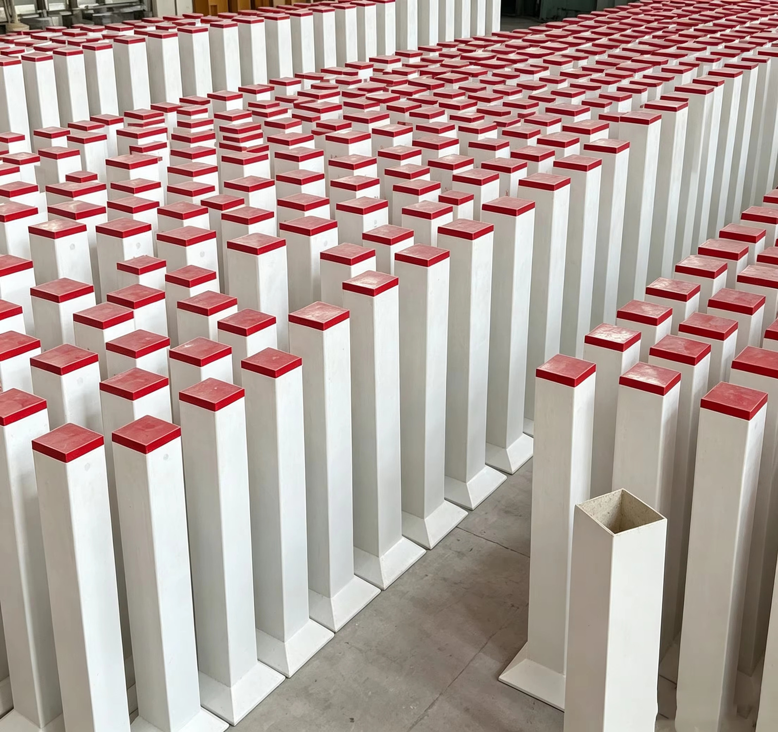 A batch of square safety warning posts with red caps laid out in rows on the workshop floor